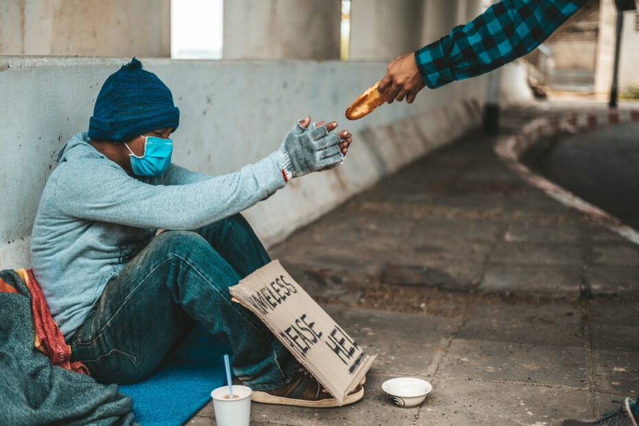 Man holding out a piece of bread to a homeless man sitting by the side of a road